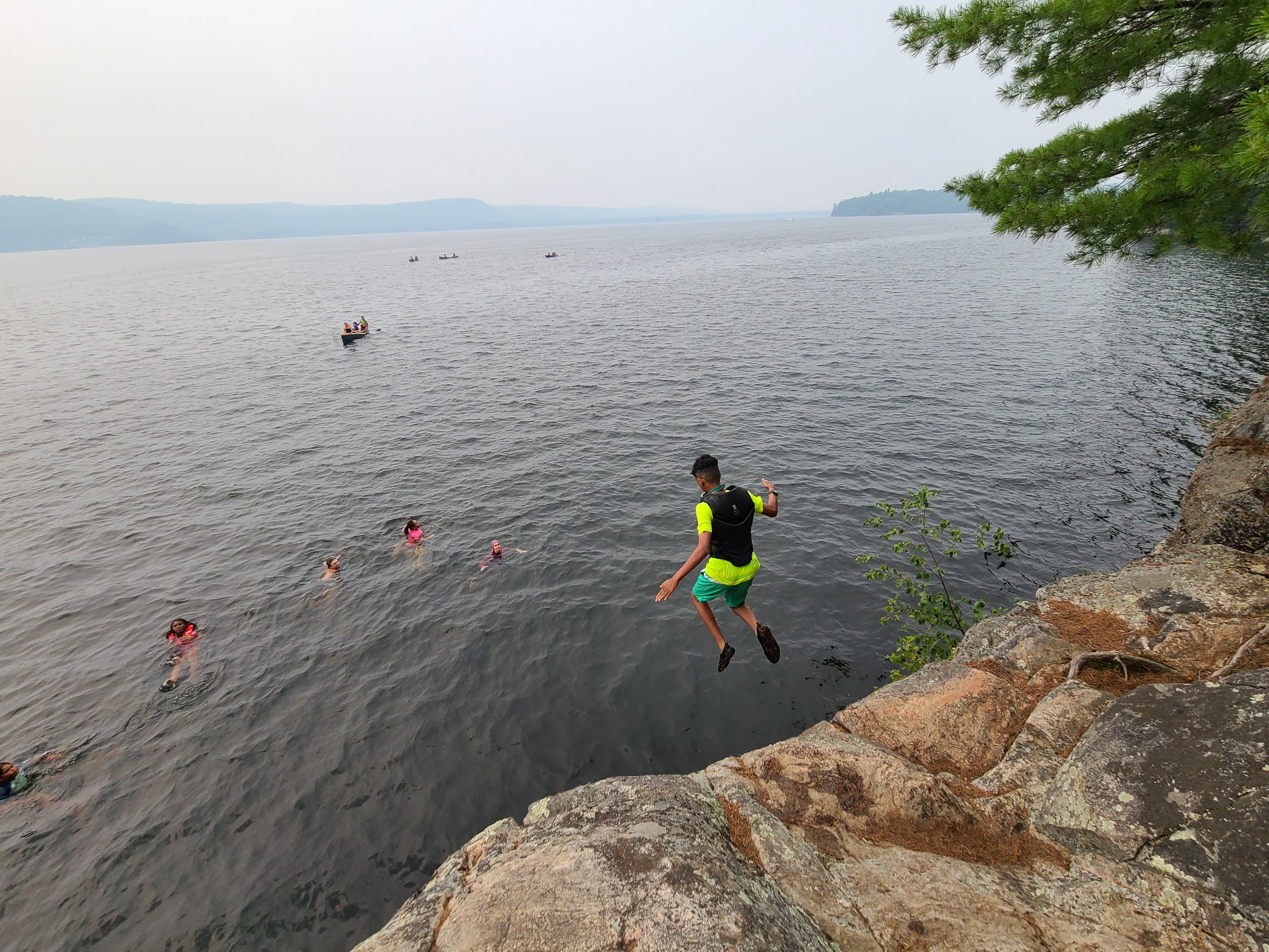 A child jumping into a lake from a rocky ledge while others swim below.